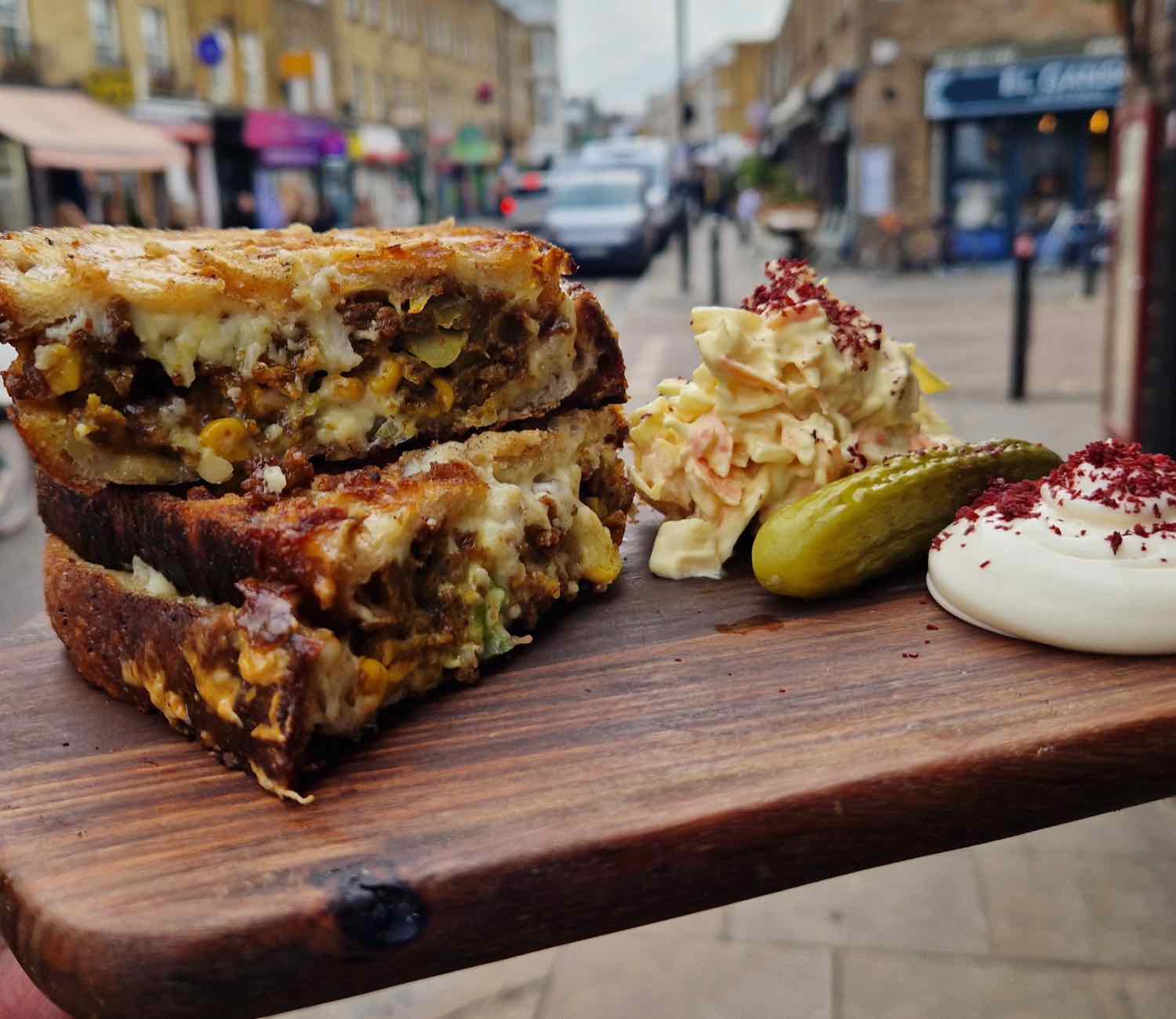 A Dalston Melts toastie cut in half with molten cheese, held up on a wooden board with Broadway Market behind