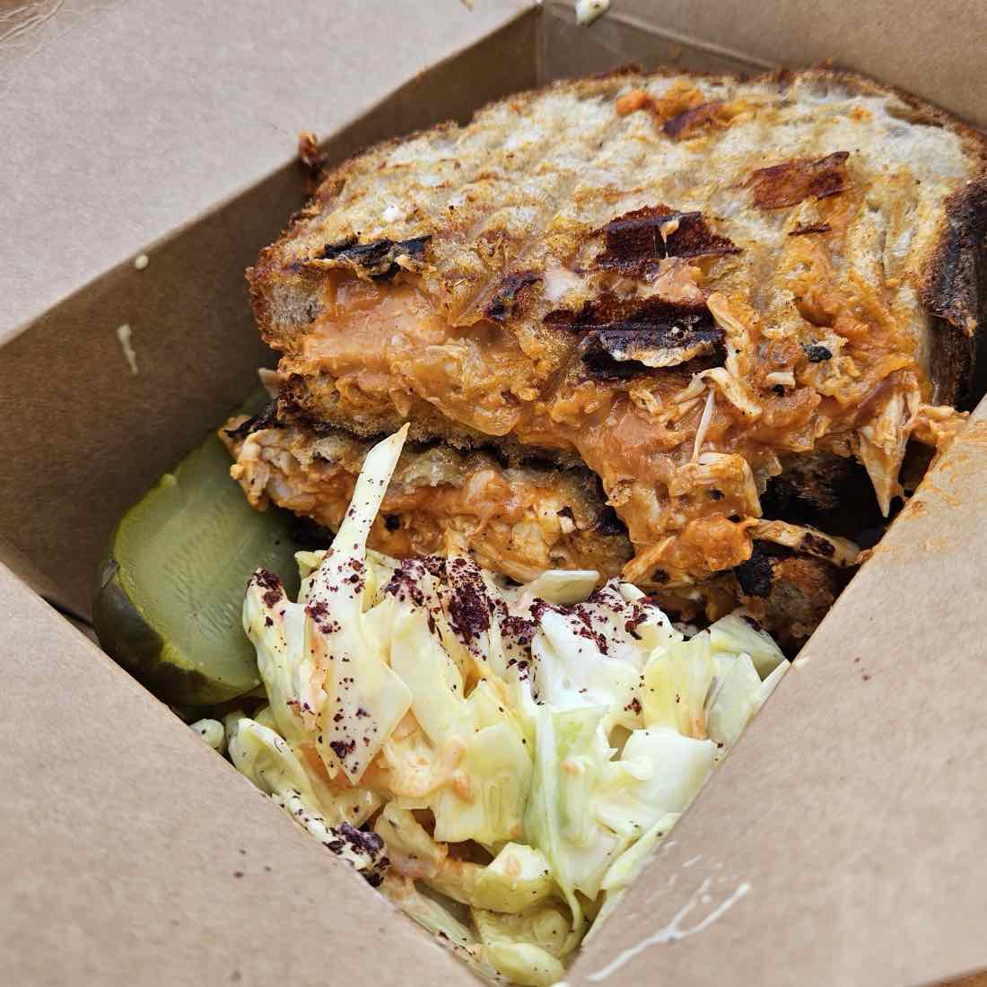 A harissa chicken melt in a kraft box with slaw and a pickle