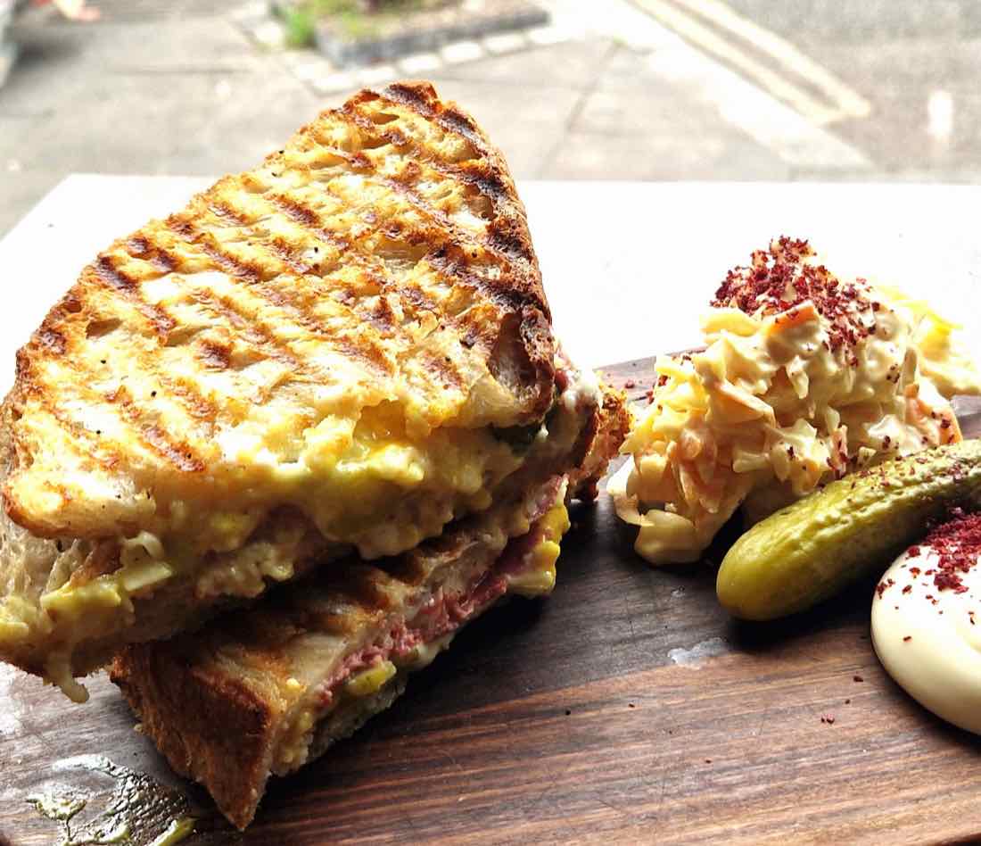 A pastrami and cheese New York Deli melt on a board with slaw, a gherkin and sour cream