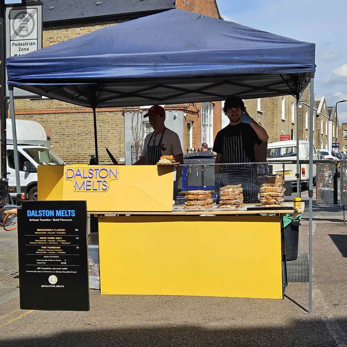 The yellow Dalston Melts market stall under a blue gazebo on Broadway Market, menu board out front and toasties stacked on the counter
